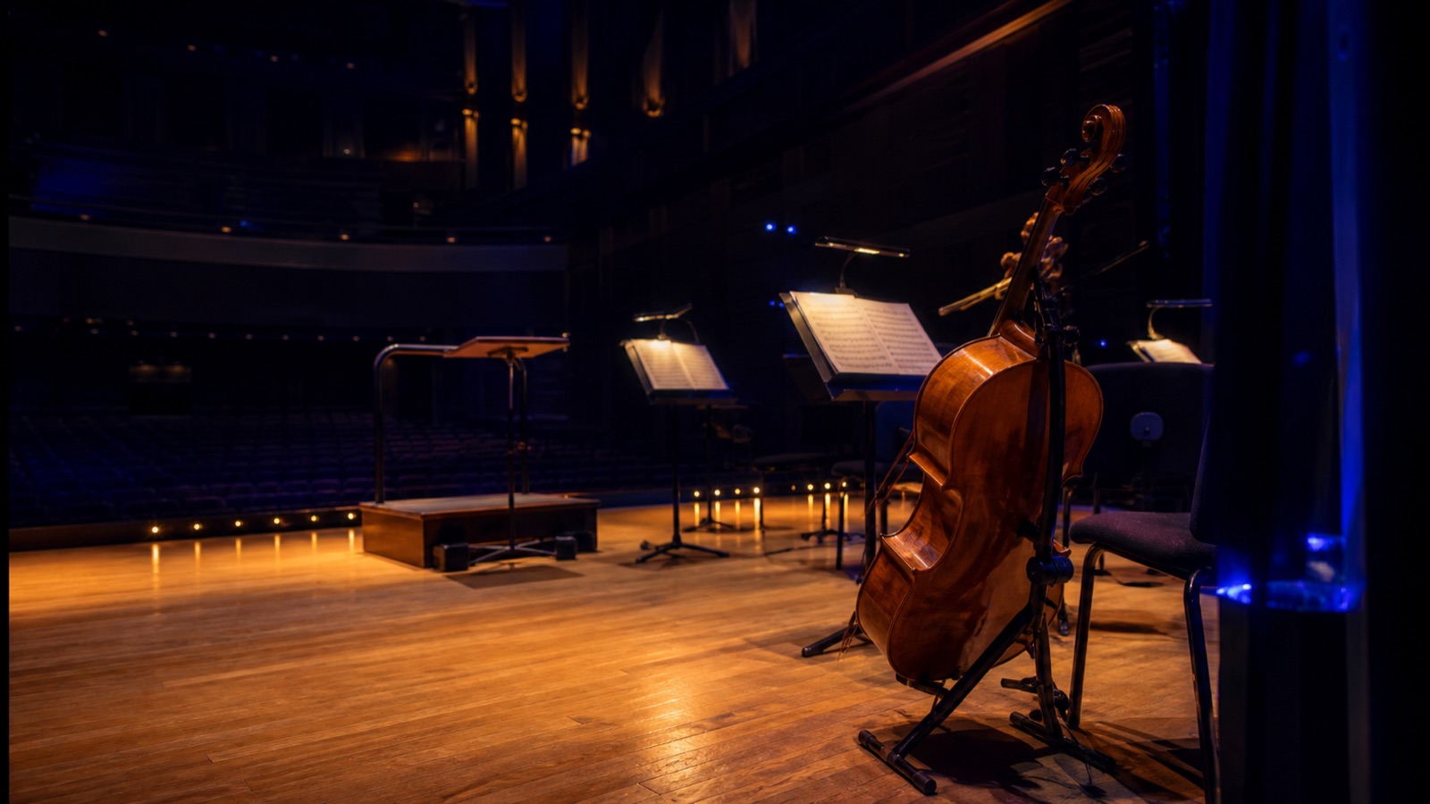 A cello and music stands wait on a warmly lit concert stage.